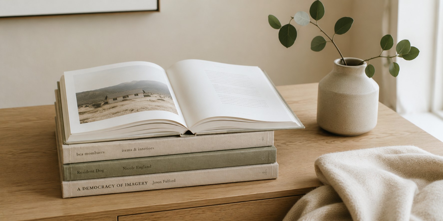 A stack of modern linen-bound books in cream and sage tones on a pale oak desk, the top book open with a sage stem bookmark, a small ceramic vessel with eucalyptus beside